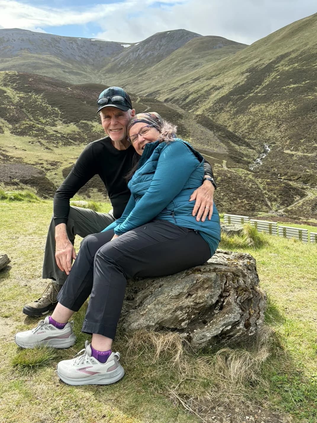 Debbie and Mike sitting together on a rock among Highland hills